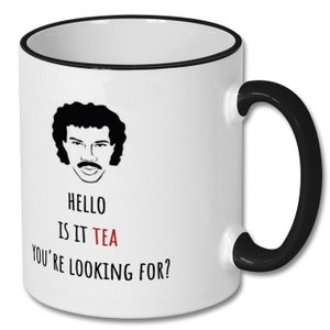May include: White ceramic mug with a black rim and handle. The mug features a black and white illustration of a man with a moustache and the text "HELLO IS IT TEA YOU'RE LOOKING FOR?" with the word "TEA" in red.