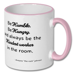 May include: White ceramic coffee mug with a pink rim and handle. The mug features a motivational quote in black text: "Be Humble. Be Hungry. and always be the Hardest worker in the room." The quote is attributed to Dwayne "The Rock" Johnson.