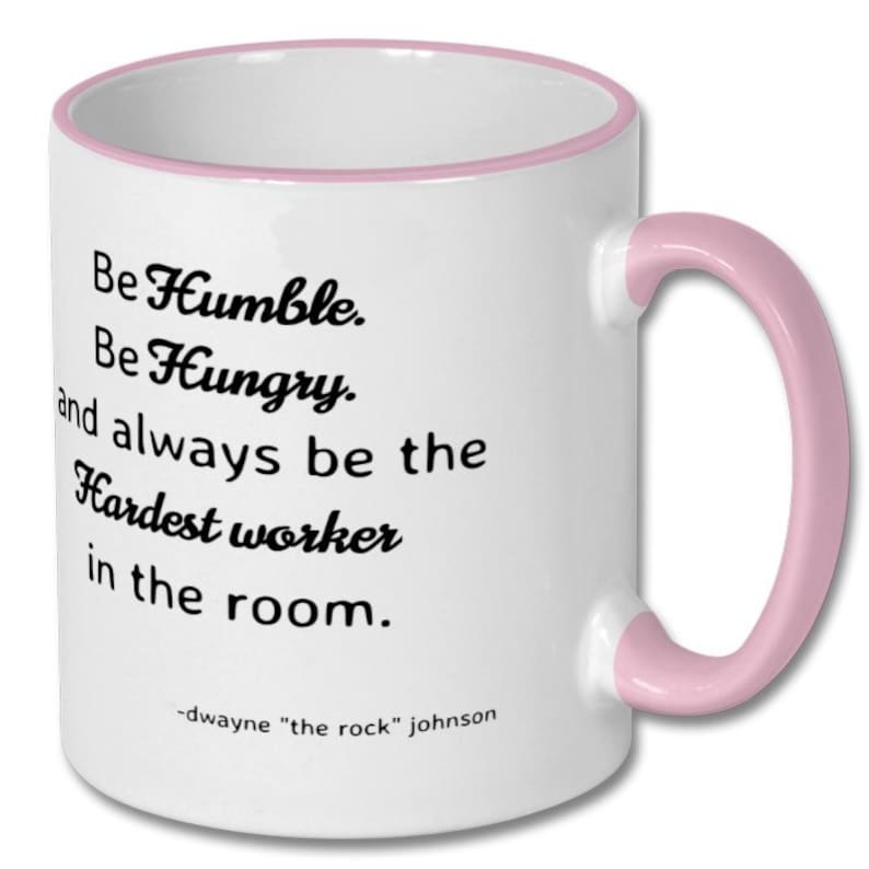 May include: White ceramic coffee mug with a pink rim and handle. The mug features a motivational quote in black text: "Be Humble. Be Hungry. and always be the Hardest worker in the room." The quote is attributed to Dwayne "The Rock" Johnson.