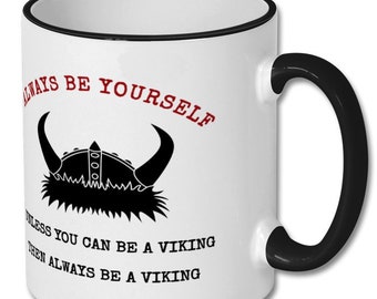 May include: White ceramic coffee mug with a black rim and handle. The mug features a black and white illustration of a Viking helmet with horns and the text "Always be yourself unless you can be a Viking then always be a Viking".