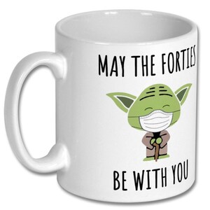 May include: White ceramic mug with a green Yoda character wearing a face mask. The text on the mug reads "May the Forties Be With You".