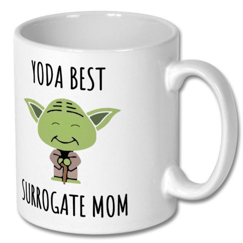 May include: White ceramic mug with a green cartoon Yoda character holding a walking stick. The text "Yoda Best Surrogate Mom" is printed on the mug.