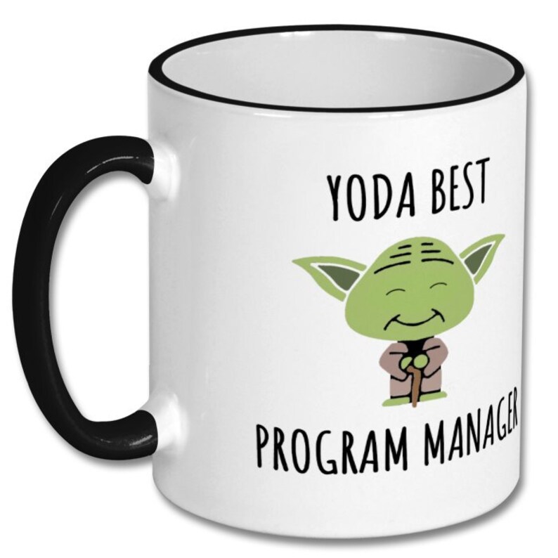 Peut inclure: Mug en c&eacute;ramique blanche avec une anse et un bord noirs. La tasse pr&eacute;sente une image de dessin anim&eacute; vert de Yoda avec le texte "YODA BEST PROGRAM MANAGER" en noir. La tasse est de taille standard.