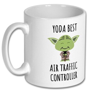May include: White ceramic mug featuring a green Yoda character illustration. The mug has black text that reads "Yoda Best Air Traffic Controller".