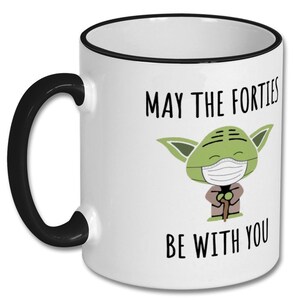 May include: White ceramic coffee mug with a black rim and handle. The mug features a green cartoon character wearing a face mask and the text "May the Forties Be With You".
