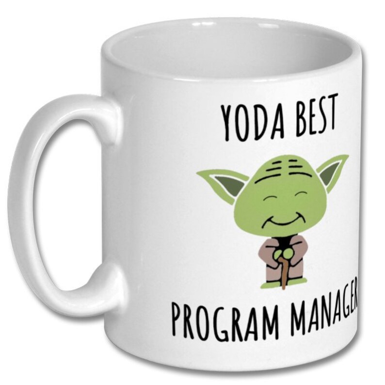 Peut inclure: Mug en c&eacute;ramique blanche avec un dessin de Yoda et le texte "YODA BEST PROGRAM MANAGER". Le personnage de Yoda est vert avec de grandes oreilles et un visage souriant. La tasse a une anse incurv&eacute;e.