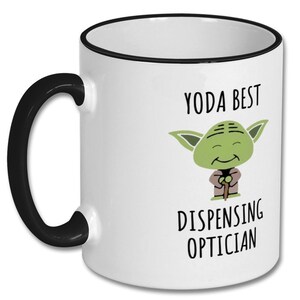 May include: White ceramic coffee mug with a black rim and handle. The mug features a cartoon image of Yoda from Star Wars with the text "Yoda Best Dispensing Optician".