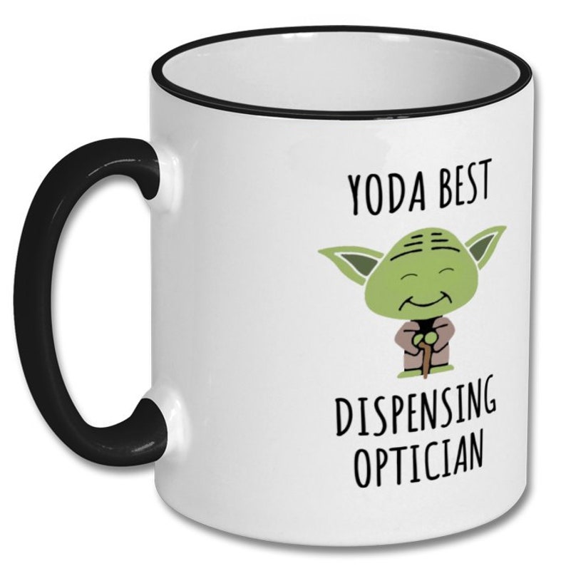 May include: White ceramic coffee mug with a black rim and handle. The mug features a cartoon image of Yoda from Star Wars with the text "Yoda Best Dispensing Optician".