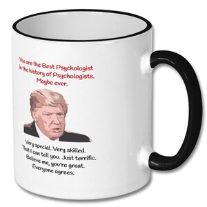 May include: White coffee mug with a black rim and handle. The mug features a cartoon image of Donald Trump and the text "You are the Best Psychologist in the history of Psychologists. Maybe ever. Very special Very skilled. That I can tell you. Just terrific. Believe me, you're great. Everyone agrees."