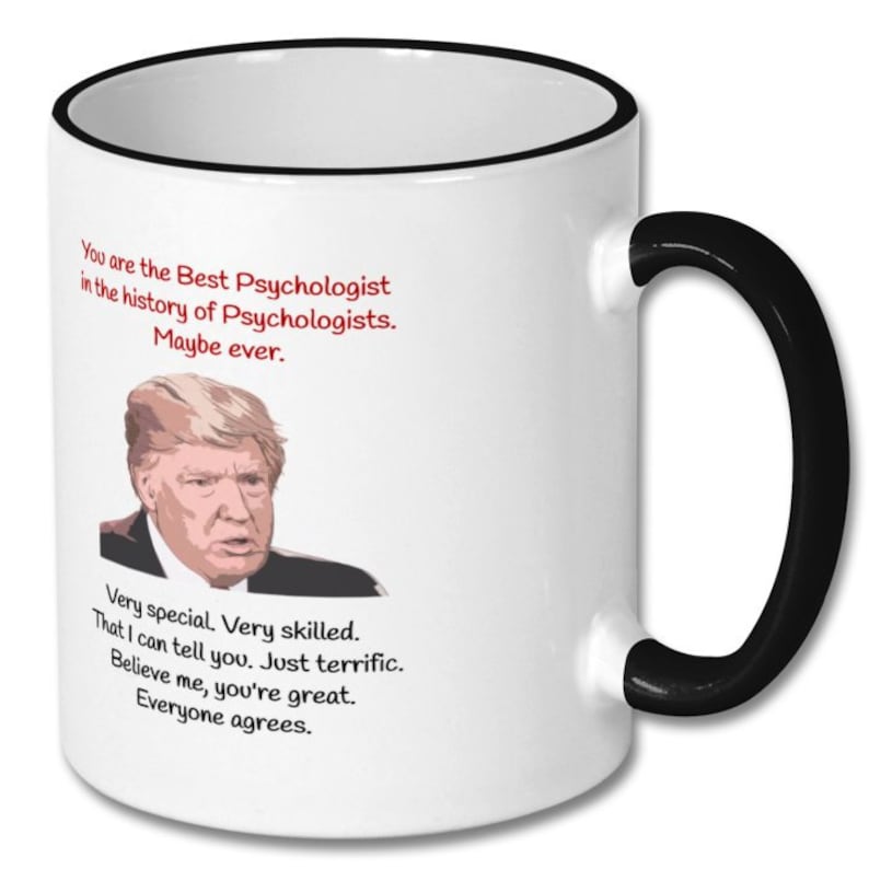 May include: White coffee mug with a black rim and handle. The mug features a cartoon image of Donald Trump and the text "You are the Best Psychologist in the history of Psychologists. Maybe ever. Very special Very skilled. That I can tell you. Just terrific. Believe me, you're great. Everyone agrees."