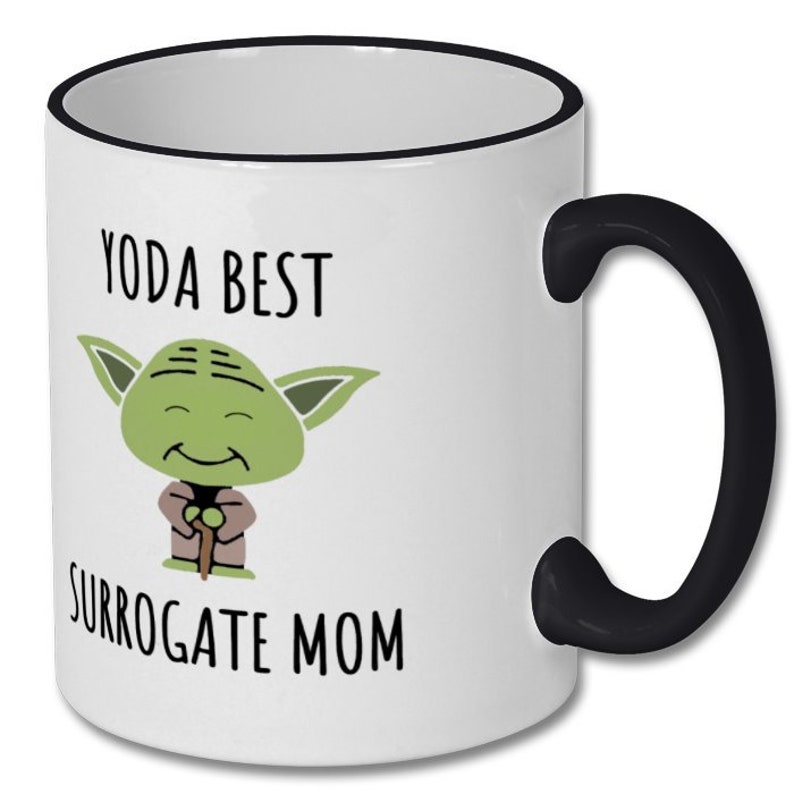 May include: White ceramic mug with a black rim and handle. The mug features a cartoon image of Yoda from Star Wars with the text "Yoda Best Surrogate Mom".