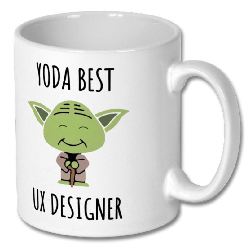 Może przedstawiać: Biały kubek ceramiczny z zieloną postacią kresk&oacute;wkową Yody trzymającą kij. Na kubku nadrukowany jest tekst "YODA BEST UX DESIGNER".