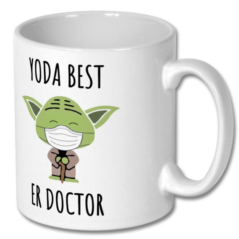 Może przedstawiać: Biały ceramiczny kubek do kawy z zieloną postacią Yody w maseczce i trzymającą laskę. Na kubku nadrukowany jest tekst "YODA BEST ER DOCTOR".