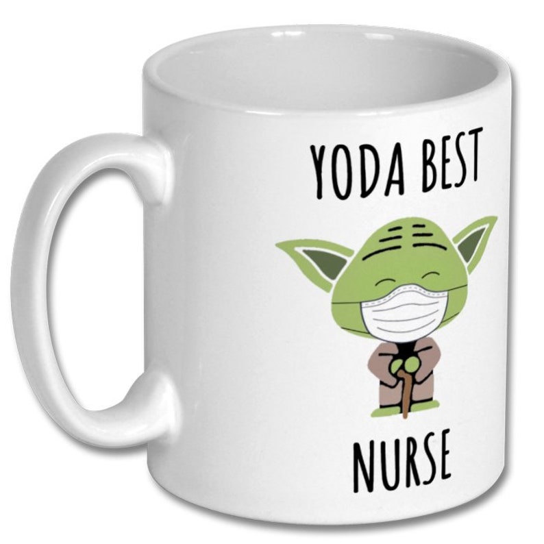 K&ouml;nnte beinhalten: Eine wei&szlig;e Keramiktasse mit einer gr&uuml;nen Yoda-Figur, die eine Gesichtsmaske tr&auml;gt und einen Stock h&auml;lt. Der Text "YODA BEST NURSE" ist auf die Tasse gedruckt.