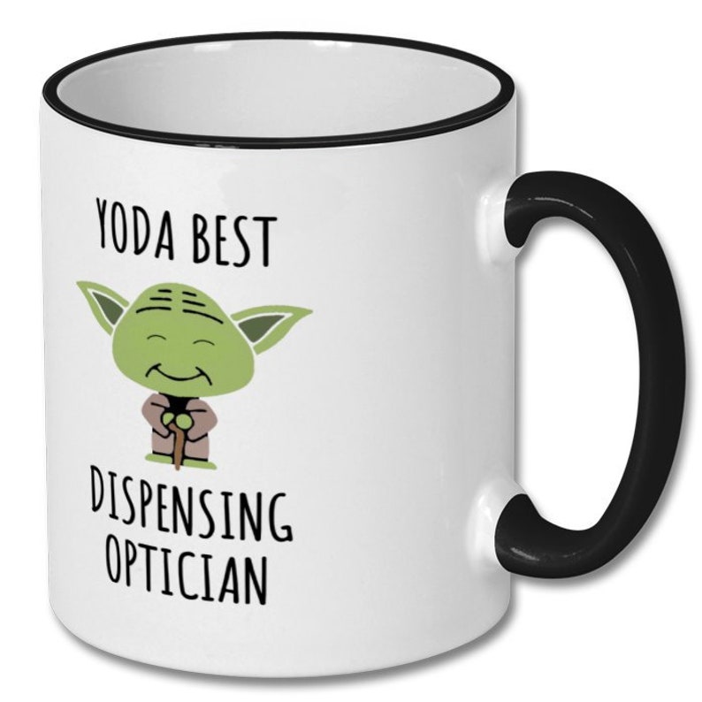 May include: White ceramic coffee mug with a black rim and handle. The mug features a cartoon image of Yoda from Star Wars with the text "Yoda Best Dispensing Optician".