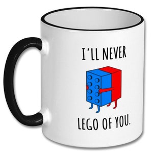 May include: White ceramic coffee mug with a black rim and handle. The mug features a cartoon illustration of two Lego blocks hugging each other. The text on the mug reads "I'll never lego of you."