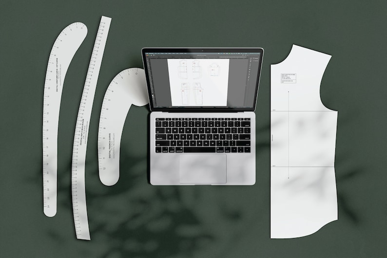 Puede incluir: Un ordenador port&aacute;til con un fondo blanco abierto a un programa de dise&ntilde;o digital. Hay tres reglas de pl&aacute;stico blanco y un patr&oacute;n de papel blanco para coser sobre una superficie verde.