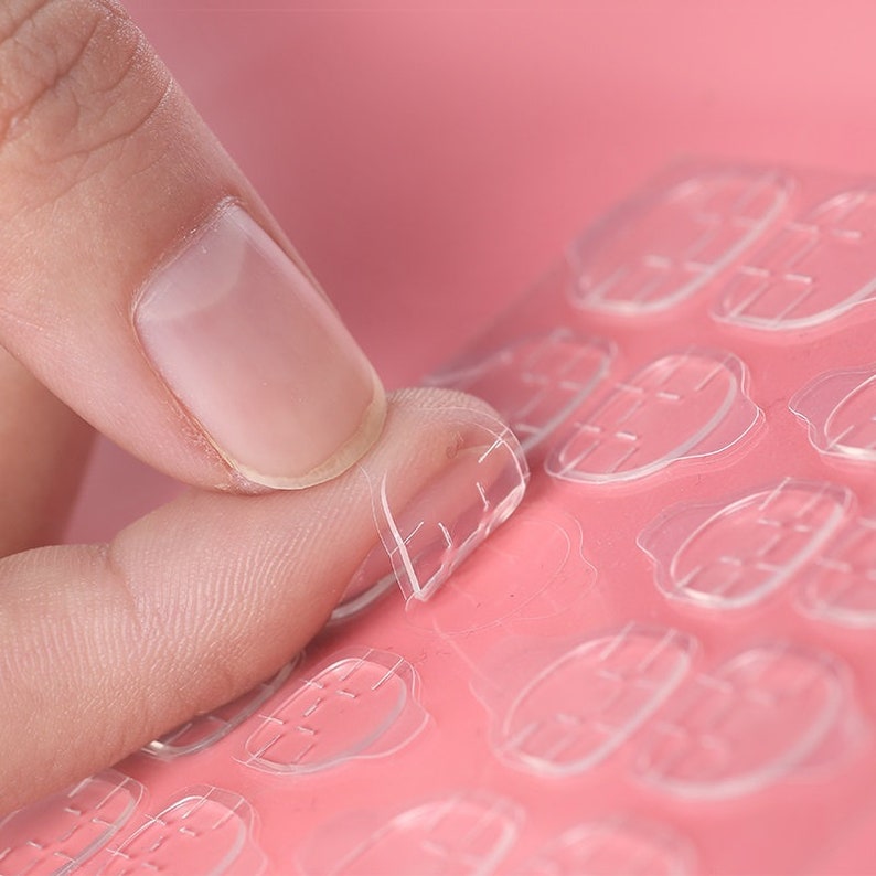 K&ouml;nnte beinhalten: Nahaufnahme von transparenten, selbstklebenden Nagel-Tabs, die von einem rosa Bogen abgezogen werden. Die ovalen Tabs haben ein Rastermuster und sind f&uuml;r das Anbringen von Kunstn&auml;geln konzipiert. Das Bild zeigt die einfache Handhabung und die Klebeeigenschaften des Produkts.