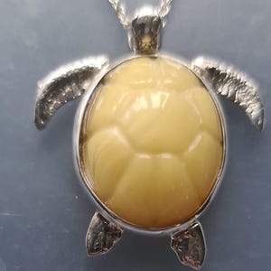 May include: A silver turtle pendant necklace. The turtle has a light yellow, textured shell and silver flipper-like arms and legs. The pendant hangs from a delicate silver chain. The background is a light blue.