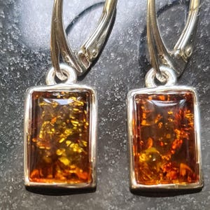 May include: A pair of rectangular amber earrings with a silver-colored metal frame and lever-back closures. The amber stones display a mix of golden and reddish-brown hues, with visible internal patterns.