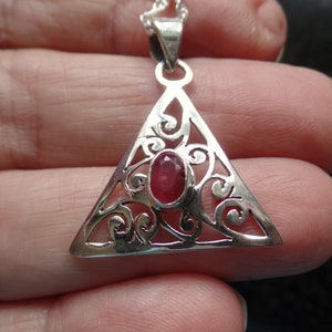 Könnte beinhalten: Eine silberne Halskette mit einem dreieckigen Anhänger, der einen roten Edelstein in der Mitte trägt. Der Anhänger hat komplizierte Filigrandetails.