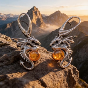 May include: A pair of silver dragon earrings with amber-colored orbs. The earrings are detailed with dragon wings and a curved tail. The earrings are displayed on a rock with a mountain range in the background.