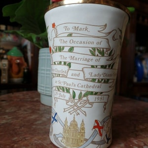 May include: White ceramic commemorative mug with gold trim, featuring a design celebrating the wedding of Prince Charles and Lady Diana Spencer at St. Paul's Cathedral on 29 July 1981. The mug depicts a stylised image of the cathedral, a ribbon with the text "To Mark The Occasion of The Marriage of", and a floral design.