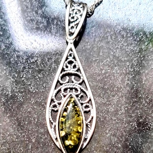May include: Silver-toned pendant necklace with a teardrop-shaped design. The pendant features an open filigree pattern surrounding a central, oval-shaped amber stone with a yellow-green hue. The necklace chain is a delicate, twisted rope style.