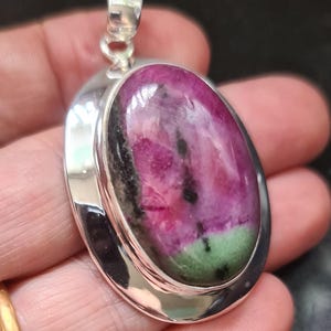 May include: A silver pendant with an oval gemstone. The gemstone is a mix of pink, green, and black. The pendant has a polished silver frame and a loop for a chain.