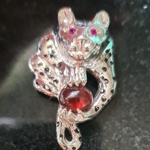 May include: A silver-toned pendant featuring a detailed sculpture of a leopard holding a red gemstone. The leopard has pink eyes and a spotted pattern.