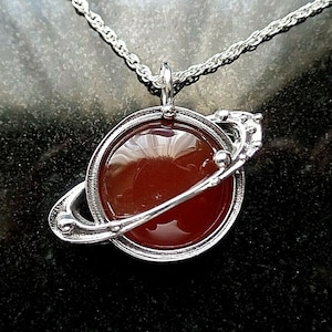 May include: A silver necklace with a pendant shaped like the planet Saturn. The pendant features a red carnelian stone in the centre.