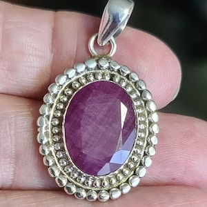 May include: Oval pendant featuring a large, faceted, deep red gemstone. The gemstone is set in a silver-coloured metal frame with a beaded border. The pendant has a silver bail for attaching to a chain.