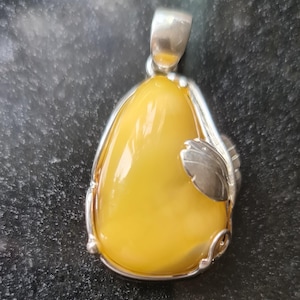 May include: A pendant featuring a large, teardrop-shaped amber stone with a silver-colored metal frame and bail. A silver leaf design is attached to the side of the amber. The amber has a glossy, yellow appearance.