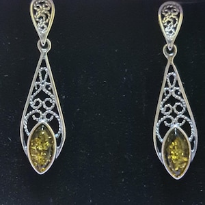 May include: A pair of silver-toned dangle earrings. Each earring features an open filigree design with a teardrop shape. The bottom of each earring has an oval-shaped amber-colored stone. The earrings are displayed against a black background.