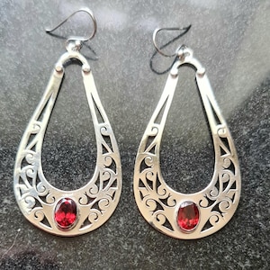 May include: A pair of silver-toned teardrop earrings with intricate openwork designs. Each earring features a vibrant red oval gemstone at the base. The earrings have a hook closure for pierced ears, and are displayed against a dark background.