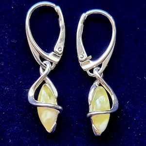 May include: A pair of silver-toned leverback earrings. Each earring features a teardrop-shaped, pale yellow gemstone encased in a decorative silver wire design. The earrings are displayed against a dark blue background.