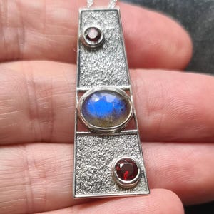 Puede incluir: Un collar con un colgante de plata con una piedra de labradorita azul y dos piedras de granate rojo. El colgante es rectangular y tiene una superficie texturizada.