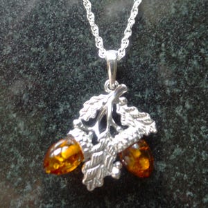 May include: Silver-toned pendant necklace featuring an oak leaf and acorn design. The pendant has two amber-colored acorn-shaped stones. The chain is a delicate silver rope style.
