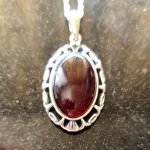 May include: Oval pendant necklace with a silver-colored filigree frame and a large, polished, dark red gemstone. The pendant hangs from a silver chain. The gemstone has a reflective surface.
