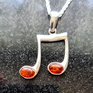 May include: A silver necklace featuring a musical eighth note pendant. The pendant has two oval-shaped amber-colored stones set within the note's design. The necklace chain is a delicate silver rope.