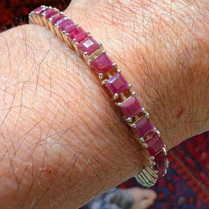 Könnte beinhalten: Ein goldfarbenes Armband mit quadratisch geschliffenen, tiefroten Edelsteinen. Die Edelsteine sind eng aneinander gesetzt und bilden eine durchgehende Farblinie. Das Armband wird am Handgelenk getragen.