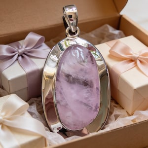 Puede incluir: Un colgante ovalado grande con una piedra preciosa de color rosa claro pulida, engastada en un marco de metal plateado liso y reflectante. El colgante está colocado en posición vertical, centrado entre tres pequeñas cajas de regalo con lazos de satén.