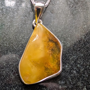 May include: A silver pendant necklace featuring a large, irregularly shaped amber stone. The amber has a warm, golden-yellow hue with darker inclusions. The pendant is set in a silver bezel and hangs from a delicate silver chain.