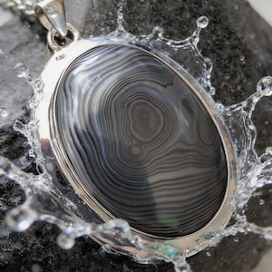 May include: Silver-framed oval pendant with a grey and white patterned stone, suspended on a silver chain. Water droplets surround the pendant, creating a splash effect. The stone displays concentric circular patterns. The pendant is a piece of jewellery.