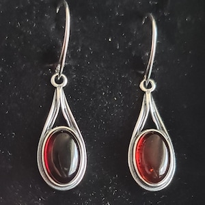 May include: A pair of silver-toned drop earrings. Each earring features a teardrop-shaped frame with a polished, oval-cut, dark red gemstone. The earrings have a simple, elegant design, suitable for everyday wear or special occasions.