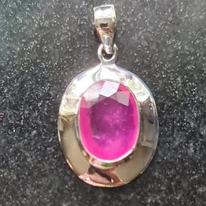 Könnte beinhalten: Silberanhänger mit einem ovalen, facettierten, magenta-farbenen Edelstein. Der Anhänger hat einen polierten Silberrahmen und eine kleine Schlaufe oben für eine Kette. Der Edelstein ist der Mittelpunkt und reflektiert das Licht.