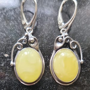 May include: A pair of silver-toned drop earrings with oval, pale yellow amber stones. The earrings have a decorative, swirling design around the amber and a lever-back closure. The background is a dark, textured surface.