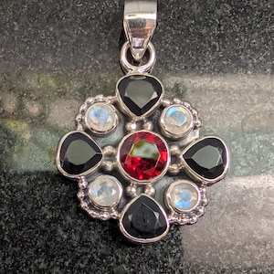 May include: A silver pendant featuring a central round red garnet surrounded by four faceted black stones and four round iridescent moonstones arranged in a floral pattern, topped with a polished silver bail for a necklace chain.