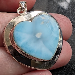 May include: A heart-shaped pendant with a light blue gemstone and white marbling, set in a silver-coloured metal frame. The pendant has a bail for attaching to a chain. The gemstone's colour is reminiscent of the ocean.