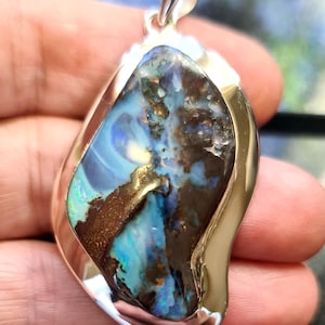 May include: A silver pendant with a striking opal gemstone. The opal showcases a blend of blue, green, and brown hues. The pendant is set in a polished, curved silver frame, with a loop for a chain.